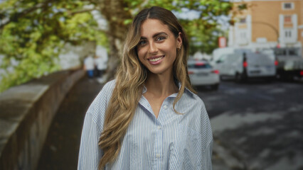 Young blonde lady face smiling with flowing hair in striped blouse on leafy sunlit sidewalk; joy...