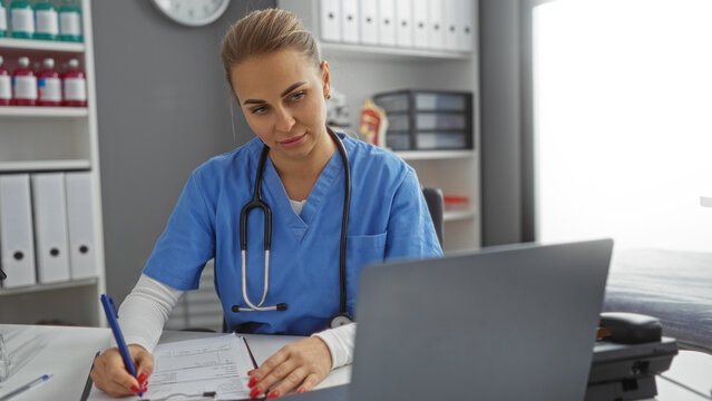 Woman doctor working at desk in hospital office, wearing blue scrubs with stethoscope, looking at laptop, writing medical notes, organized shelves in clean interior setting