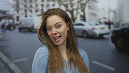 Young brunette woman in blue sweater sticks tongue out on busy street amid moving cars; playful...