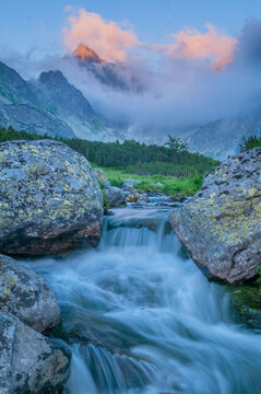 Fototapeta View of a vibrant river flowing through lichen-covered boulders towards misty mountains touched by the warm glow of sunrise, VysokÃ© Tatry, PreÅ¡ov Region, Slovakia.