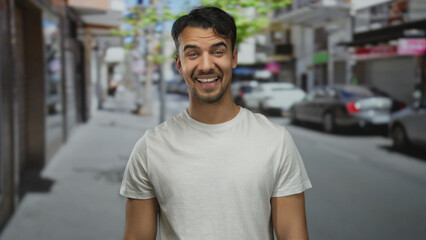 Young hispanic man smiling on city street wearing casual white shirt surrounded by urban environment with cars and shops in the background on a sunny day outdoors