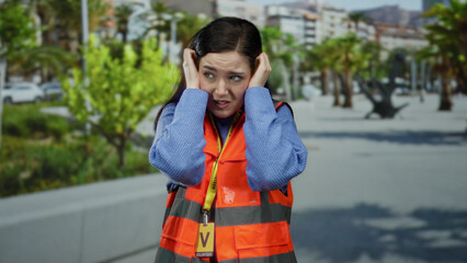 Obraz premium Woman in orange vest covers ears from noise on busy street with green trees in the background, expressing discomfort outdoors.