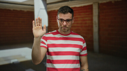 Young man with glasses waving at a construction site, wearing a red striped shirt, exhibiting...
