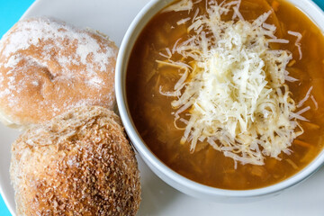A Bowl Of Traditional French Onion Soup Topped With Grated Cheese And White Bread Rolls Topped With Flour And Seeds