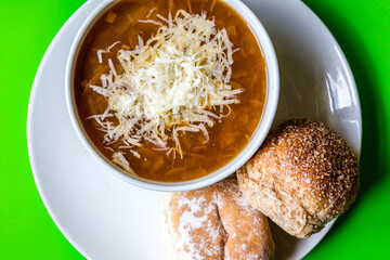 A Bowl Of Traditional French Onion Soup Topped With Grated Cheese And White Bread Rolls Topped With Flour And Seeds