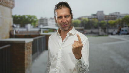 Blond man with long hair shows middle finger gesture with bare hand on street by stone railing;...