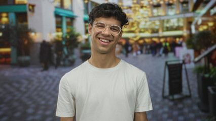 Man with curly hair and glasses turns his head to the side and smiles on a crowded city street,...