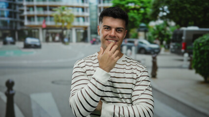 Fototapeta premium Young man standing on a city street in a striped shirt, smiling warmly in an urban outdoor environment with blurred background of vehicles and buildings.