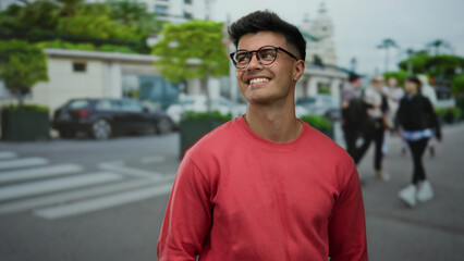 Young man in red sweater smiles confidently outdoors in an urban city street with lush greenery and...