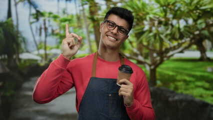 Young man in glasses, wearing red and apron, holding coffee cup, stands smiling and pointing...