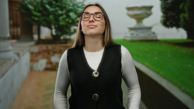 Woman standing outdoors with closed eyes in peaceful urban garden setting wearing glasses and casual vest on a sunny day