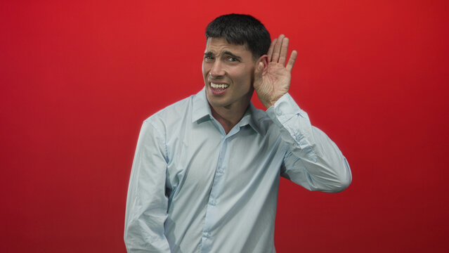 Man holds hand to ear listening against red backdrop in studio with inquisitive expression; curiosity.