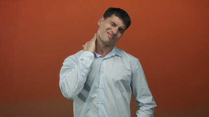 Young hispanic man wearing light shirt holds neck in studio as he grimaces with pain against orange...