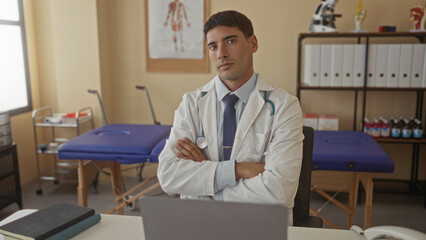 Man doctor wearing white coat and tie with stethoscope and crossed arms at desk in building;...