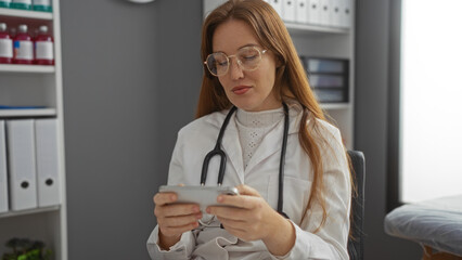 Woman in clinic wearing glasses and lab coat sitting with smartphone looks focused in hospital...