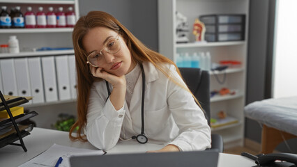 Woman doctor with red hair looks thoughtful in a clinical setting, surrounded by medical equipment and paperwork in a hospital room.