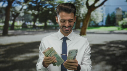 Fototapeta premium Young hispanic man in white shirt counting dirham banknotes with hands in forest clearing; prosperity.