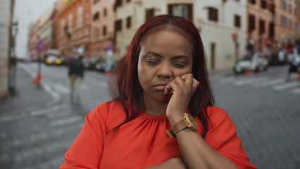 Woman resting cheek on hand wearing orange top and wooden watch on a busy city street lined with...