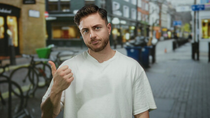 Fototapeta premium Young man with beard in white shirt giving thumbs up on city street surrounded by shops and bicycles outdoors.