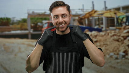 Young man with beard smiling and pointing to his smile wearing an apron and gloves at a...