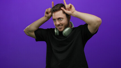 Young man with beard wearing headphones makes playful horn gesture against purple wall in isolated...