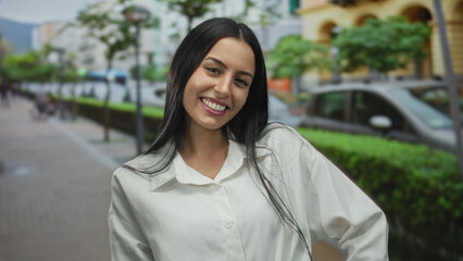 Woman smiling outdoors in a city street; young hispanic woman with long hair and white shirt enjoying urban environment on a sunny day.