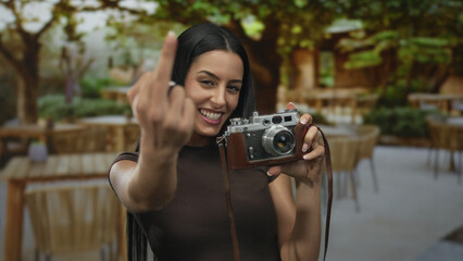 Woman holding camera on restaurant terrace outdoors making unexpected gesture with middle finger...