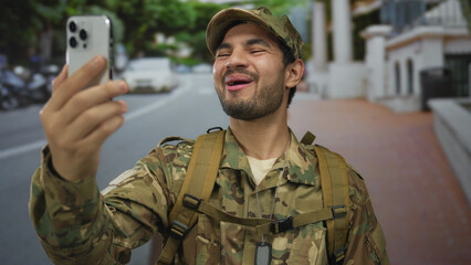 Man in camouflage uniform holds smartphone at arm's length on street under daylight; pride duty patriotism camaraderie.
