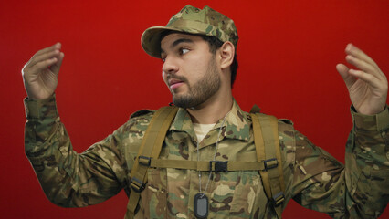 Man wearing camouflage uniform, backpack straps and dogtags gestures with both raised hands in...
