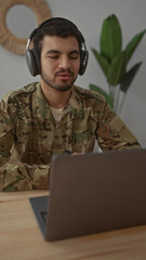 Man in military uniform listening with headphones at laptop in a cozy modern living room interior;...
