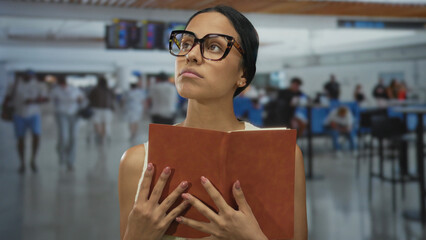 Young woman in glasses engrossed in reading a book while standing in a bustling airport terminal with people in the background, suggesting travel or waiting.