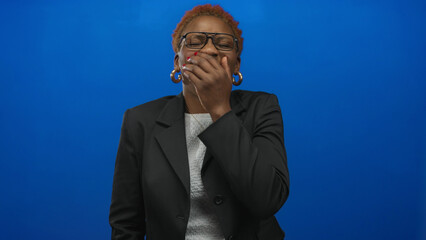 Young african american woman with glasses and hoop earrings covers mouth with hand in studio;...