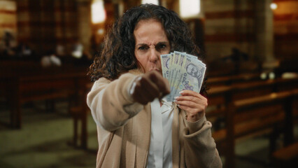Middle-aged woman holds romanian banknotes indoors, making a gesture with her fist in a church setting, expressing defiance or determination.
