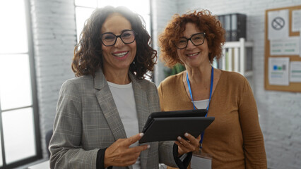 Women collaborating in an office setting as business partners, sharing ideas and smiling while engaged in work indoors with a digital tablet, showcasing teamwork and professionalism.