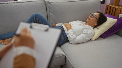 Woman lying on sofa in living room with clipboard in foreground, suggesting therapy or consultation setting, capturing mature indoor relaxation scene.
