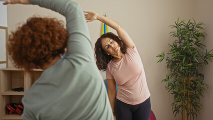 Women stretching in gym together showing friendship and fitness in an indoor sports center setting with middle-aged and hispanic elements in a peaceful atmosphere.