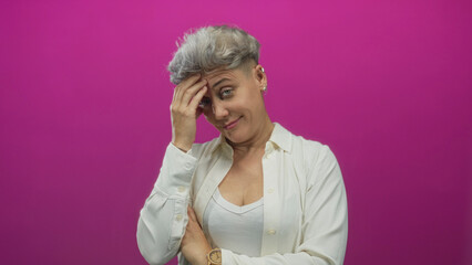 Woman with short hair with hand on forehead in pink studio holding her other arm folded across...