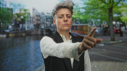 Woman with short hair wearing vest and white shirt points finger in street canal scene near river...