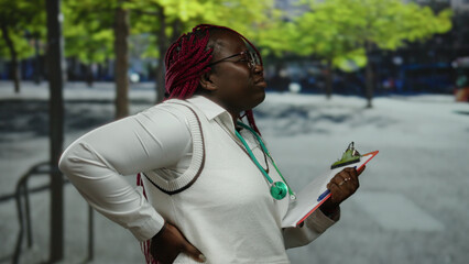 Doctor woman standing on a city street holding a clipboard and experiencing back pain with trees in...