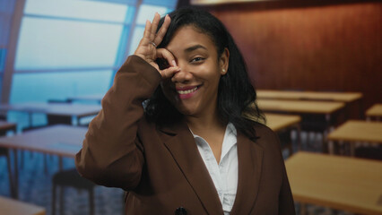 Woman in brown blazer and white shirt smiling and making ok sign with hand to eye in classroom;...