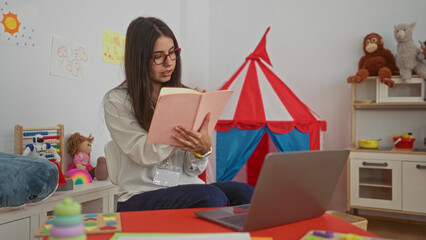 Hispanic woman teacher engaged in online teaching from a colorful kindergarten classroom filled...