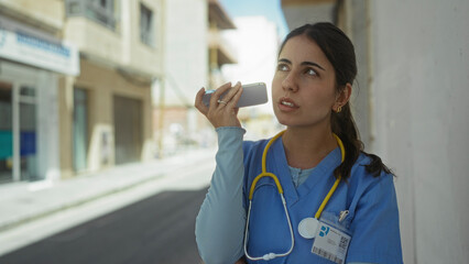 Young woman doctor in blue scrubs holds smartphone to her ear on a sunny street with buildings...