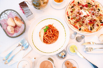 Outdoor brunch flat lay with gourmet Italian food. Table set-up with seafood pasta, Capricciosa pizza, bread basket, butter, cup of coffee, pot of tea