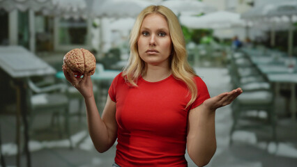 Woman in restaurant holding brain wearing red shirt with confused expression, surrounded by tables under white umbrellas. © Krakenimages.com