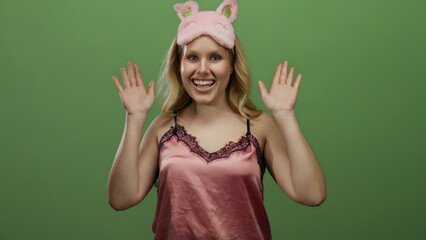 Woman in pink nightwear with a playful eye mask poses against a green background, radiating joy and...