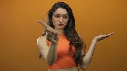 Young woman with wavy brown hair presenting hand and pointing finger in an orange studio setting;...