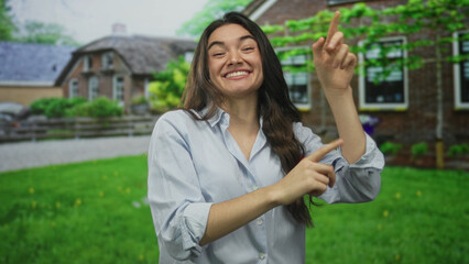 Woman with bright smile pointing finger at brick house entrance on street in green lawn outdoors;...