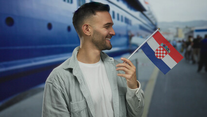 Young man holding croatian flag happily at a port with ships in the background.