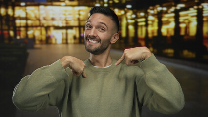 Fototapeta premium Young man pointing at himself and smiling on an urban city street with blurred lights in the background showcasing a lively outdoor atmosphere.
