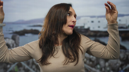 Young hispanic woman uses hand talking gesture on seaside promenade overlooking ocean;...